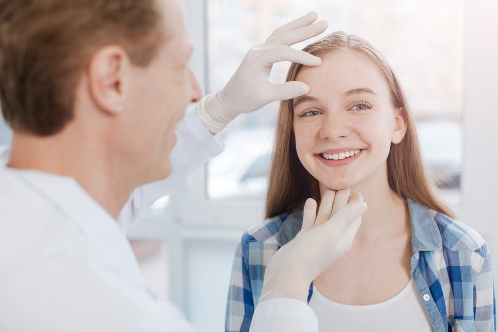 Médico branco analisando o rosto de uma paciente mulher, jovem e branca que está sorrindo.