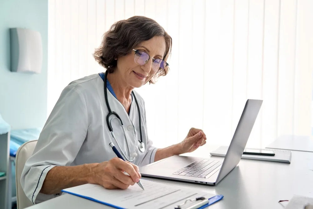 Médica sentada em sua mesa e realizando anotações em sua prancheta.