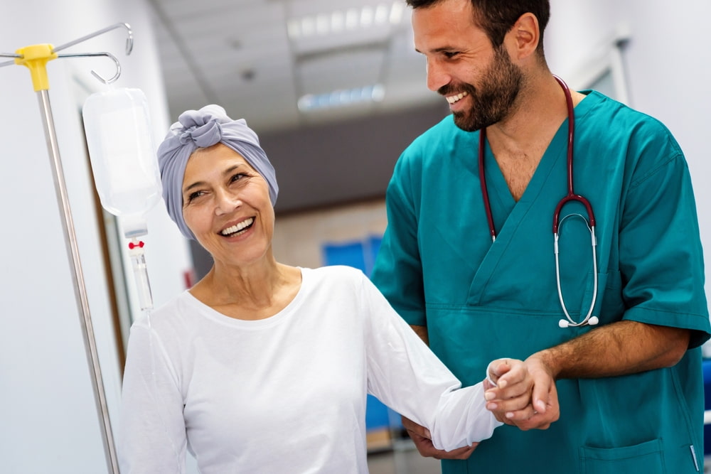 Médico auxiliando paciente oncológica a andar, enquanto ela sorri.